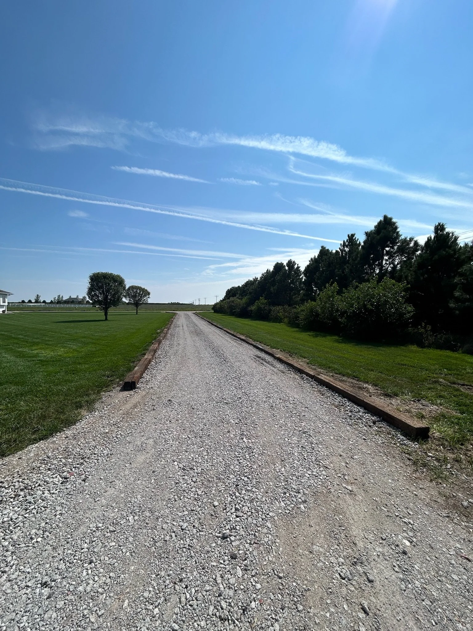 Gravel road in a rural landscape with green grass, trees, and a clear blue sky with wispy clouds.