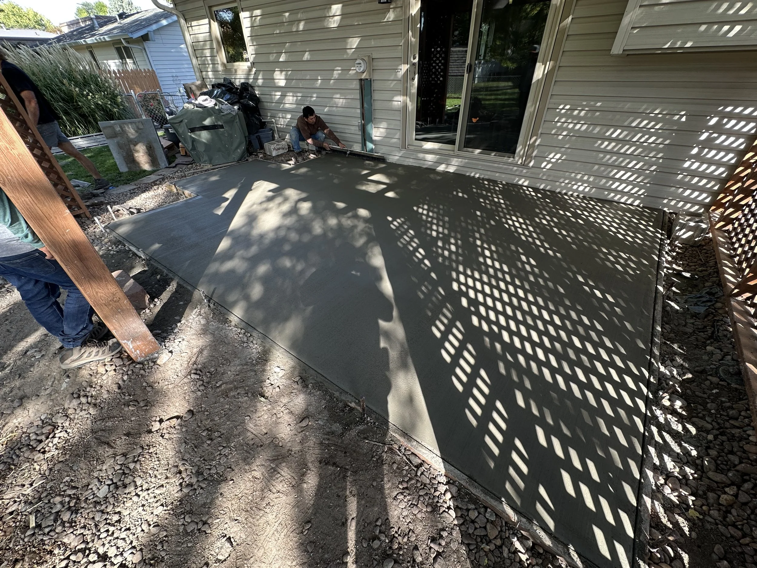 Workers installing fresh concrete on a patio area, adjacent to a house. Shadows from lattice structure create patterns on the concrete surface.