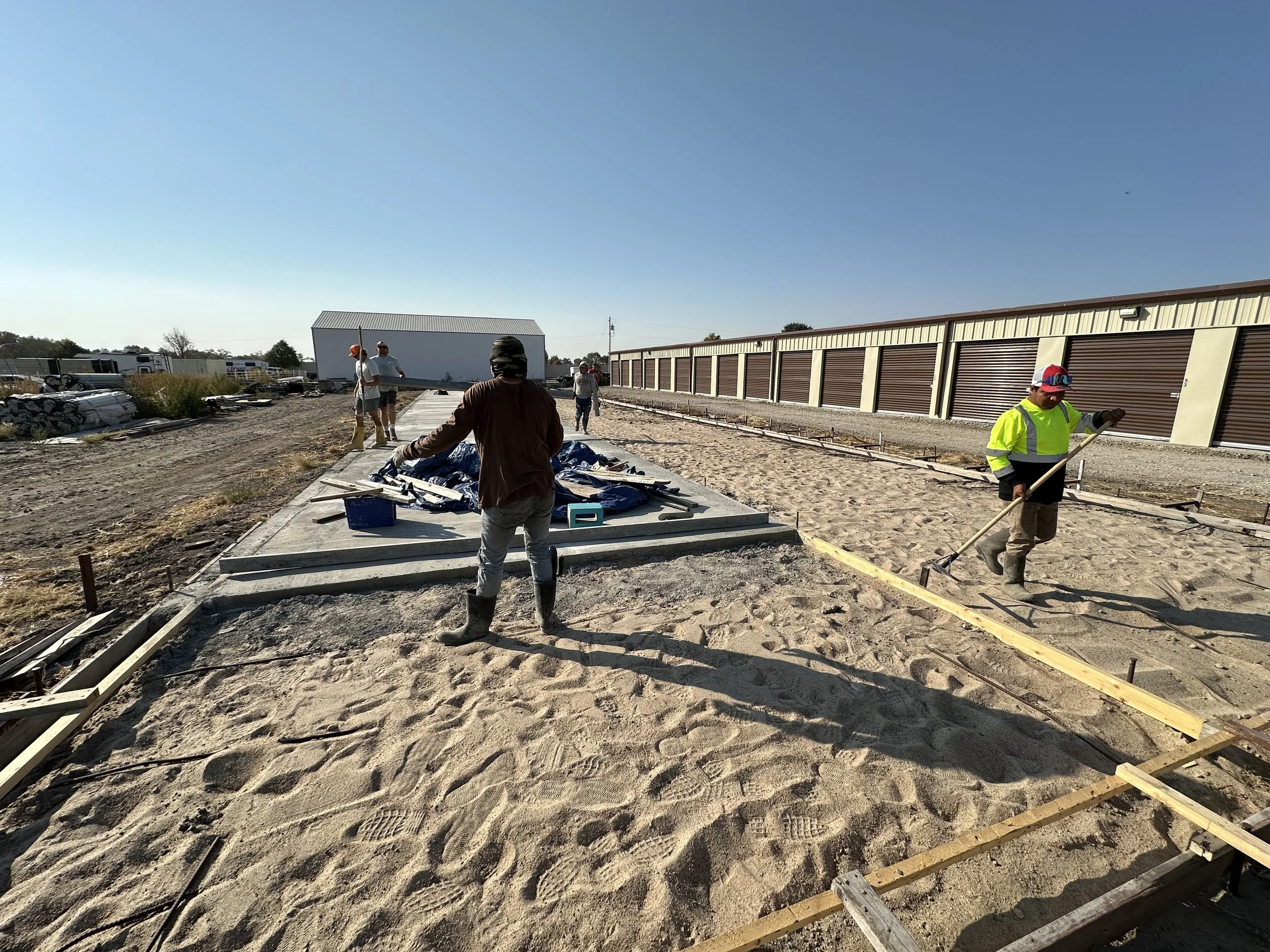 Construction workers preparing a site, laying sand on a concrete base with surrounding storage units.