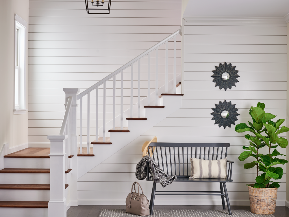 Entryway with white shiplap walls, hardwood flooring, a black bench with a pillow and coat, green houseplant in a woven basket, two decorative wall mirrors, and a staircase with white railing and dark wood steps.