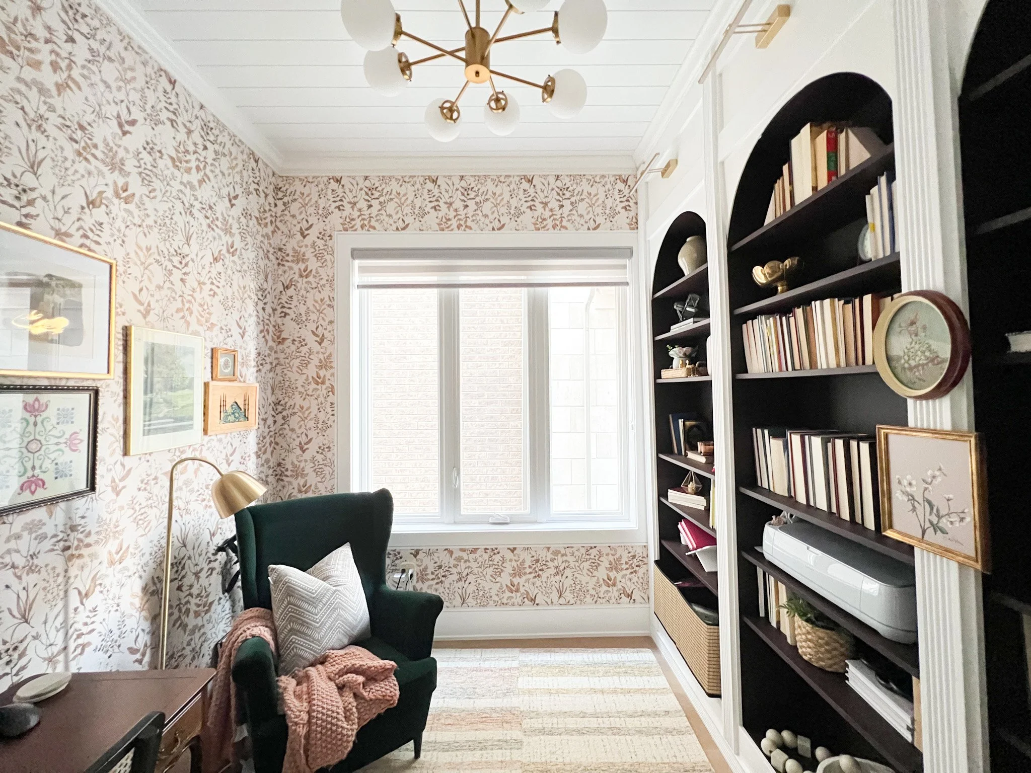 A cozy home office or reading nook with a dark green armchair, an armchair blanket, a white pillow, a gold desk lamp, white built-in bookshelves filled with books and decorative items, a window with white shades, and a ceiling light fixture with roun
