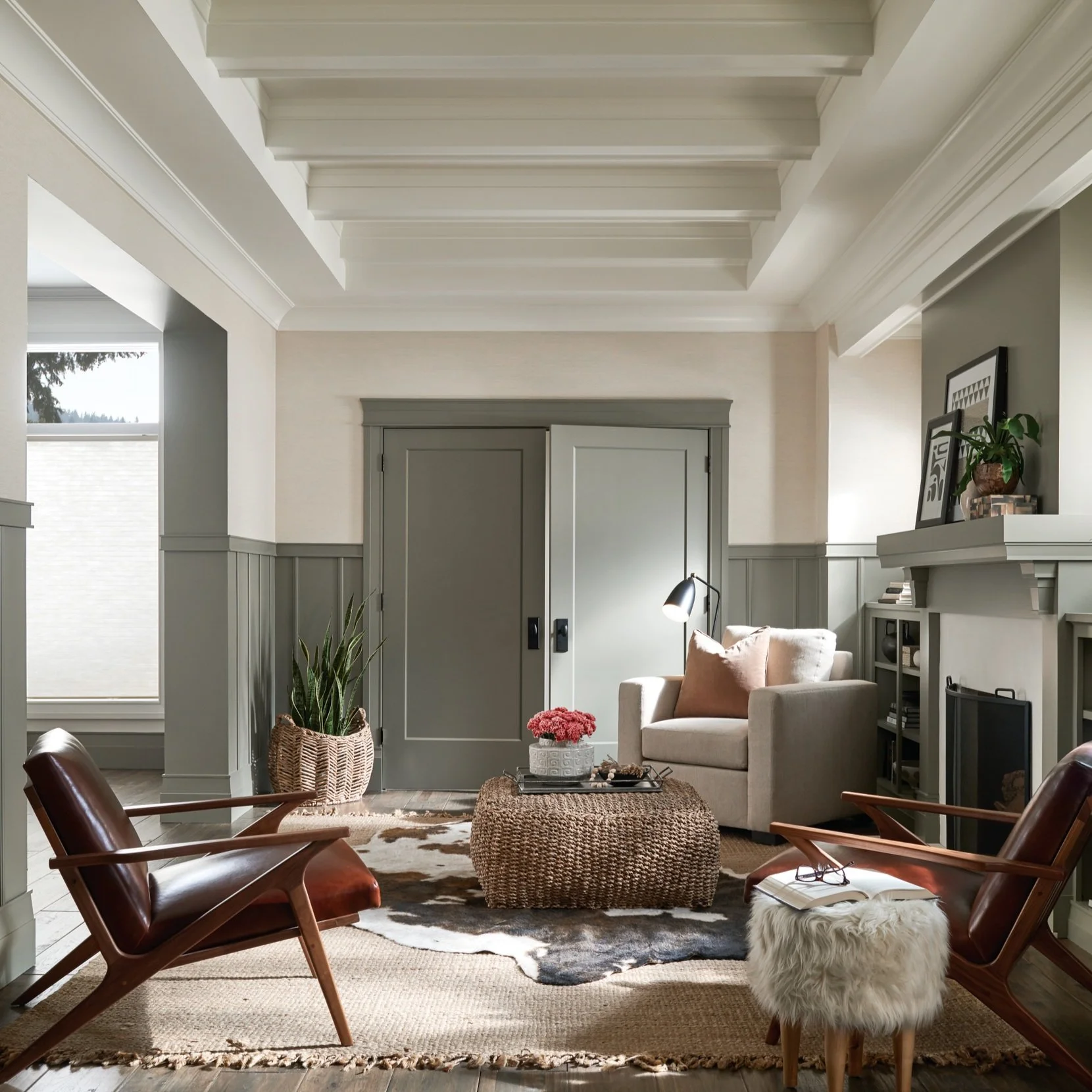 Living room with beige armchair, two wooden chairs, woven ottoman, cowhide rug, and fireplace with decor.