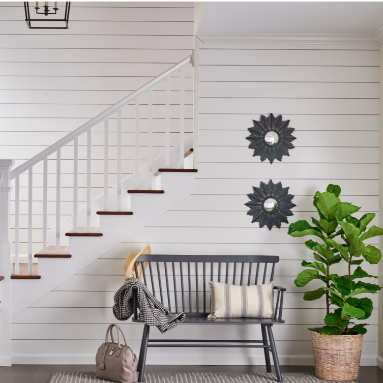 A modern hallway with white shiplap walls, a staircase with white risers and dark wood treads, two decorative black sunburst mirrors on the wall, a black bench with a striped pillow, a beige bag on the floor, a scarf draped over a hat on the bench, and a large potted fiddle leaf fig plant.