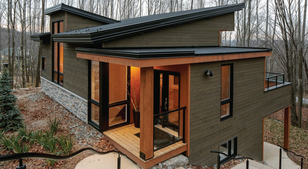 Modern multi-story house with dark exterior siding, large windows, and a small front porch with wooden flooring and railing, surrounded by trees.
