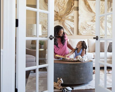 A woman in a pink shirt and a young girl are sitting at a round table in a living room, looking at each other and smiling. The woman is showing the girl a book. The room has beige walls, abstract art on the wall, and French doors with glass panes.