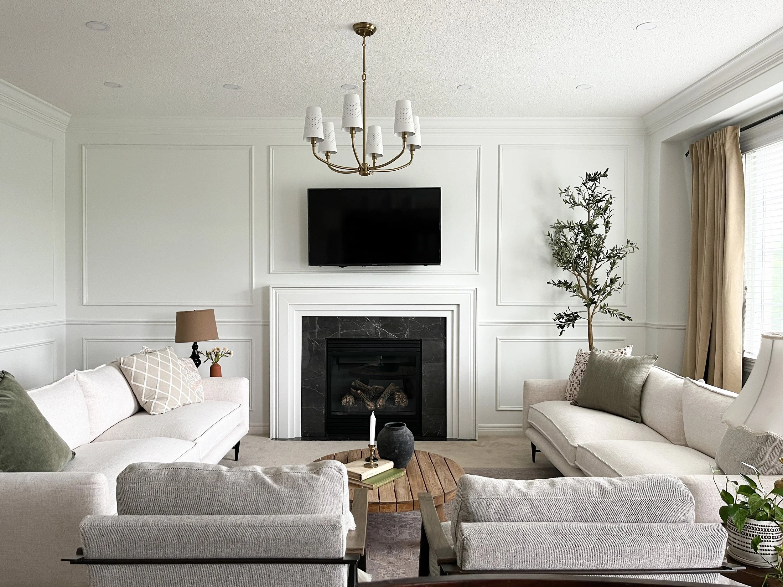 Living room with white sofas, a coffee table, fireplace, flat-screen TV, and a window with beige curtains.