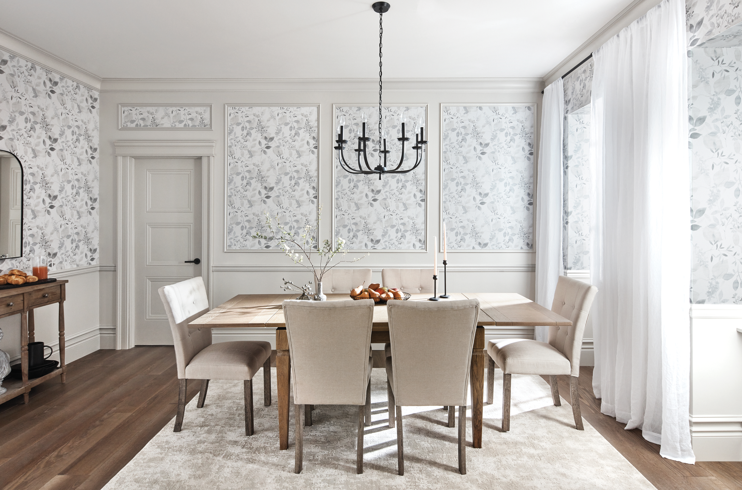 A bright dining room with a wooden table, six upholstered chairs, a black chandelier, and white curtains. Decor includes a branch with flowers and a fruit bowl on the table, with floral patterned wallpaper on the walls.