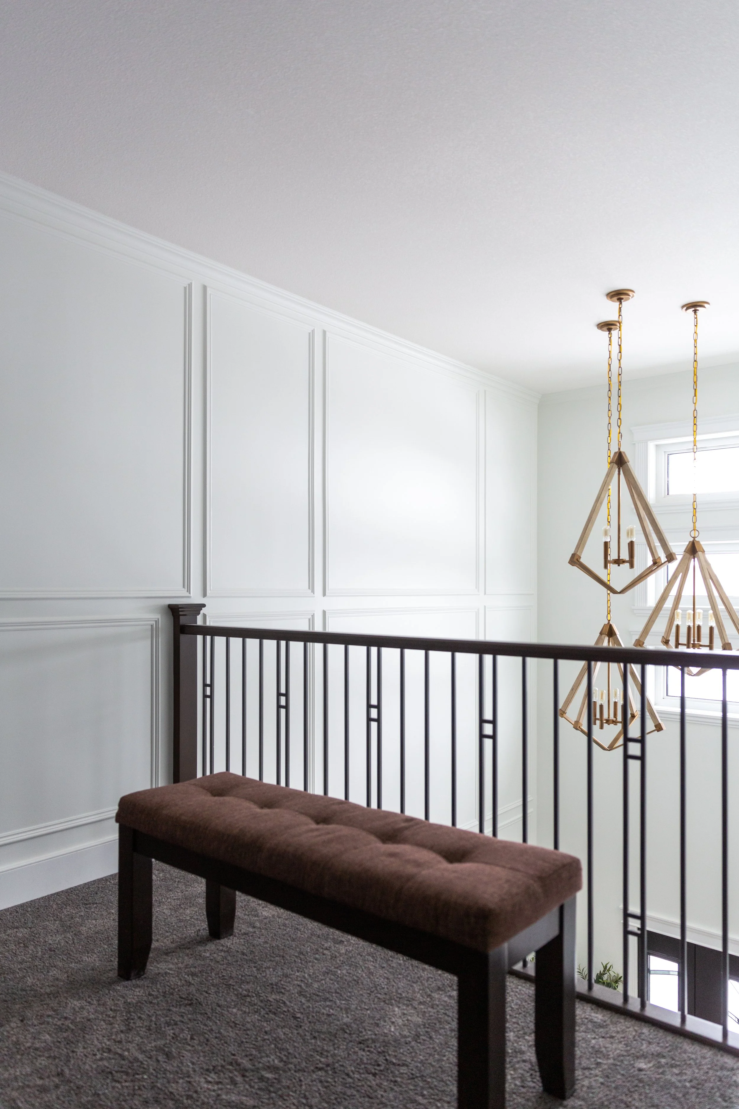 A well-lit upstairs landing area featuring a brown cushioned bench, white paneled walls, a black metal railing, and geometric chandeliers hanging from the ceiling near a window.