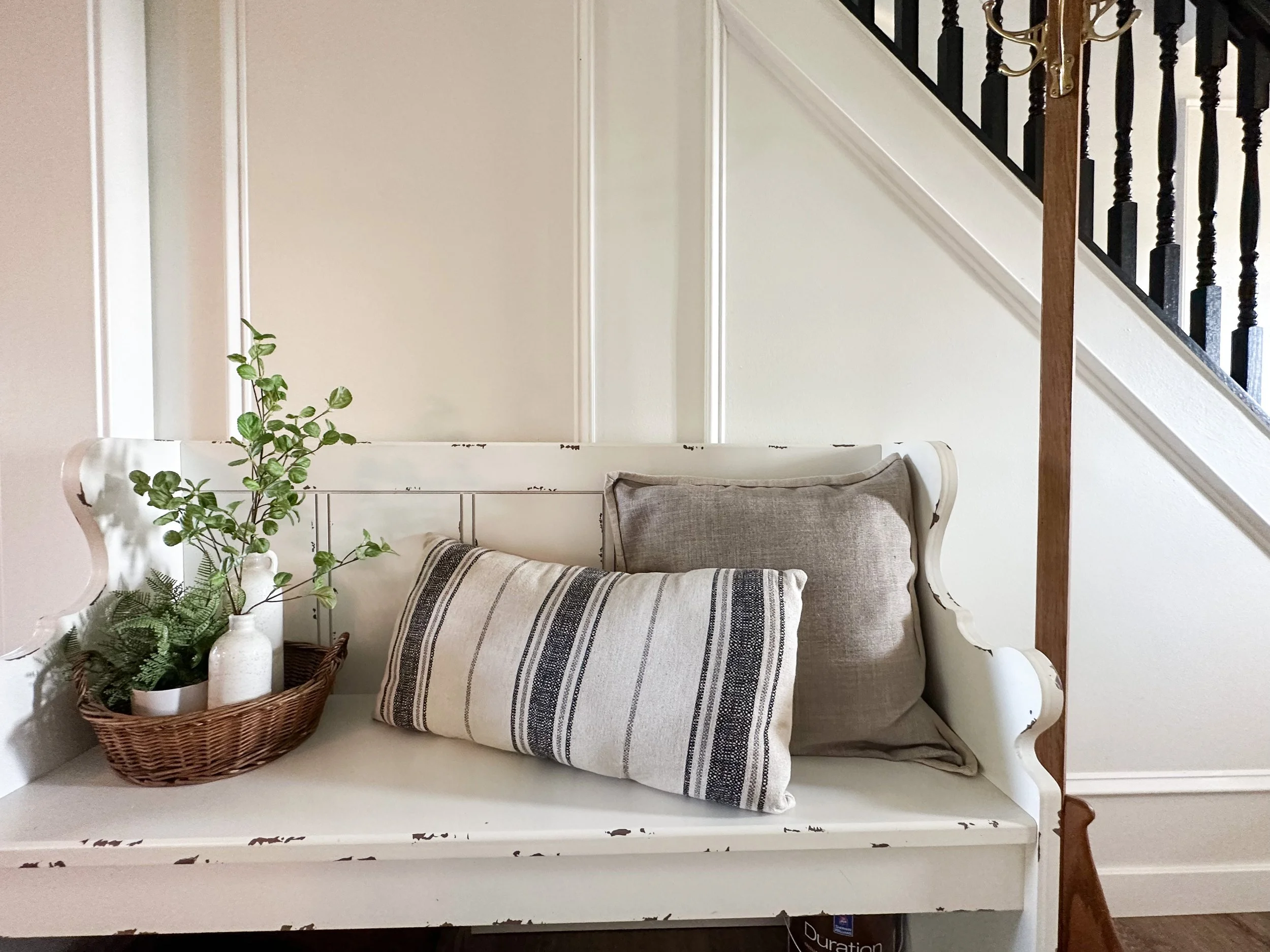 White wooden bench with distressed finish holding a wicker basket with green plants, and two pillows, one in a plain beige fabric and the other striped in black and white. The corner of a staircase with dark spindles and a brown wooden railing is vis