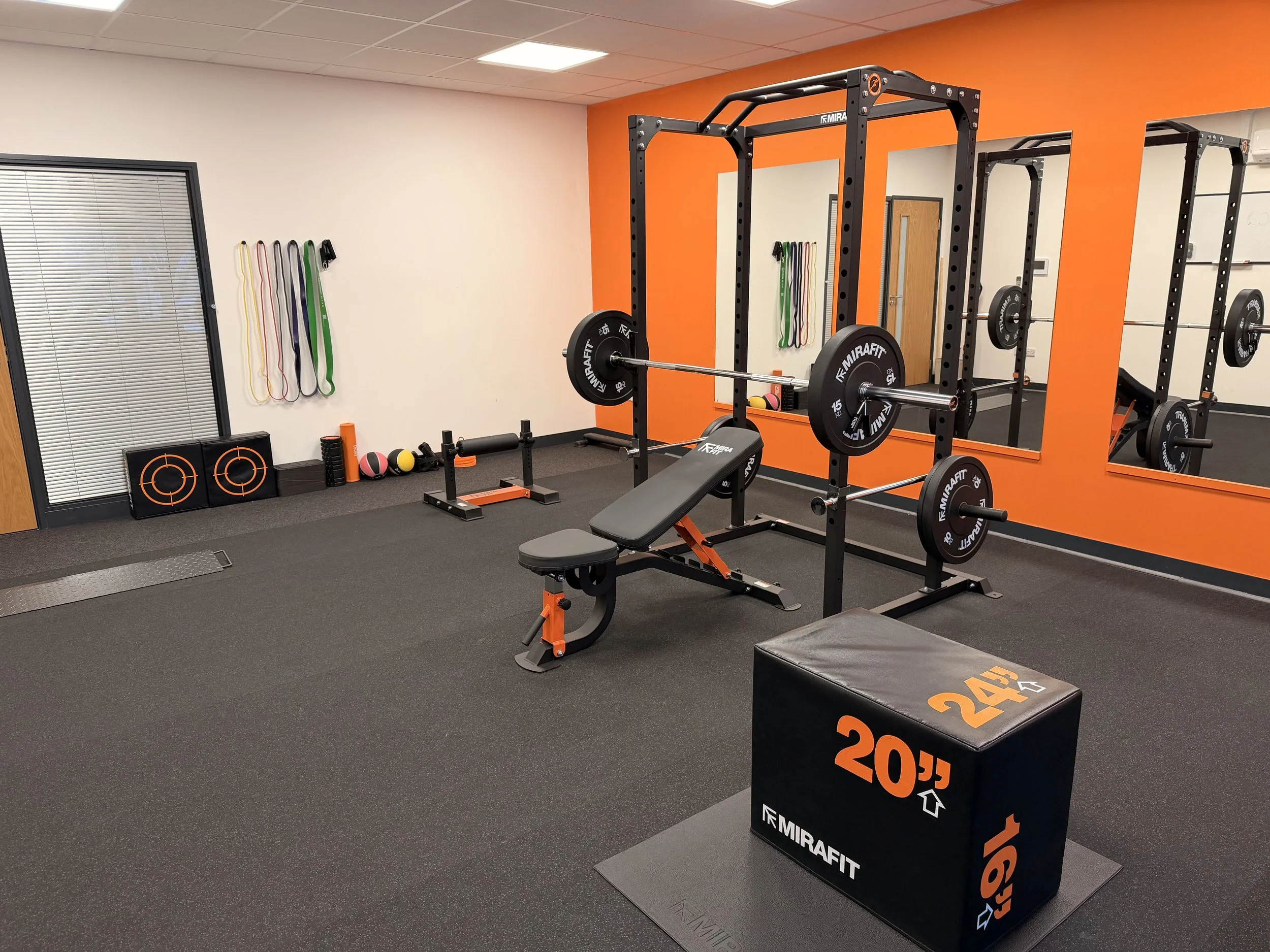 Empty gym with weightlifting equipment including a barbell on a squat rack, a workout bench, a large box with weight measurements, mirrors, and wall-mounted resistance bands.