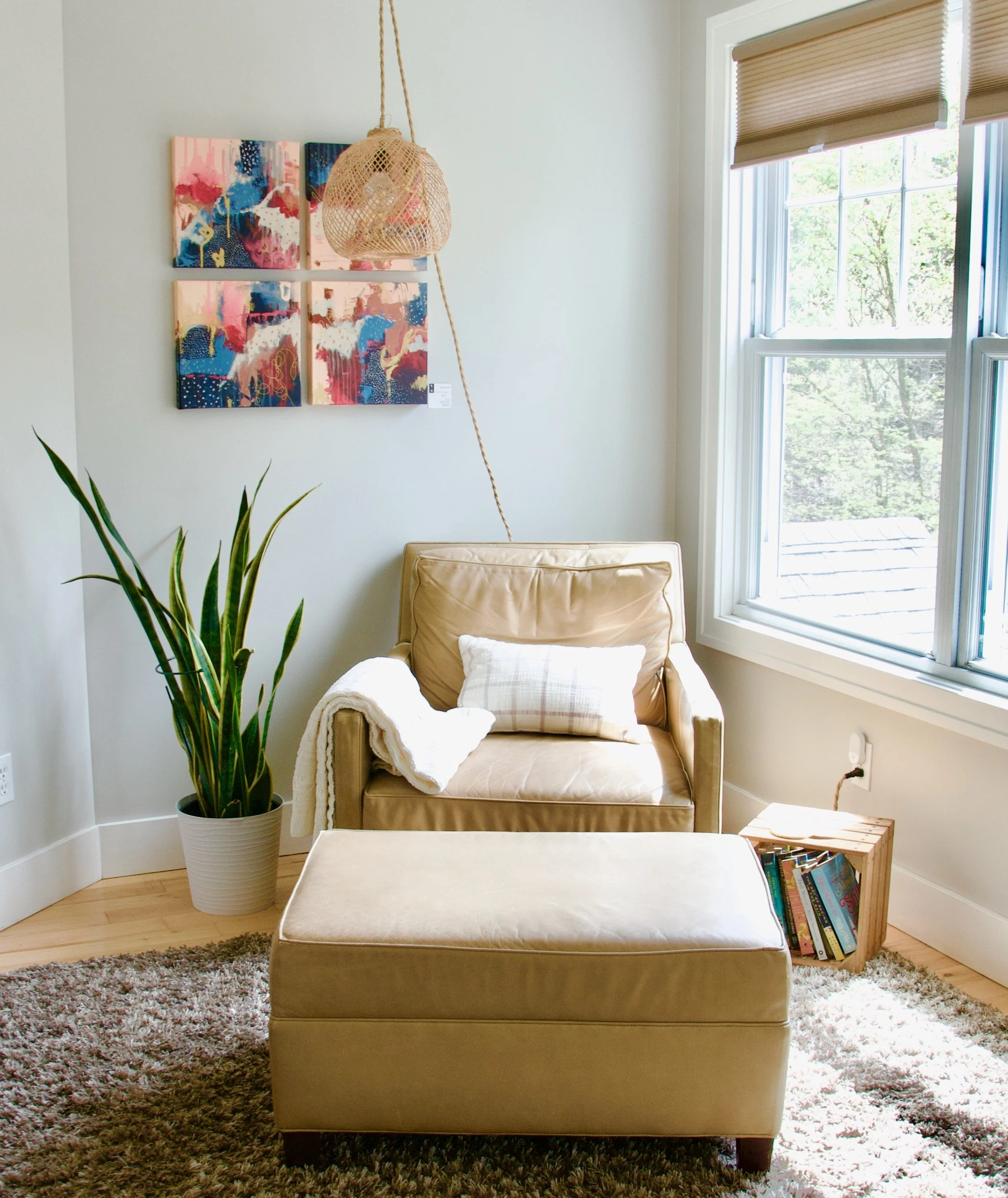 Living room corner with beige leather chair and ottoman, large plant in white pot, window with blinds, abstract art on white wall, hanging woven pendant light, small bookshelf, and a plush rug.