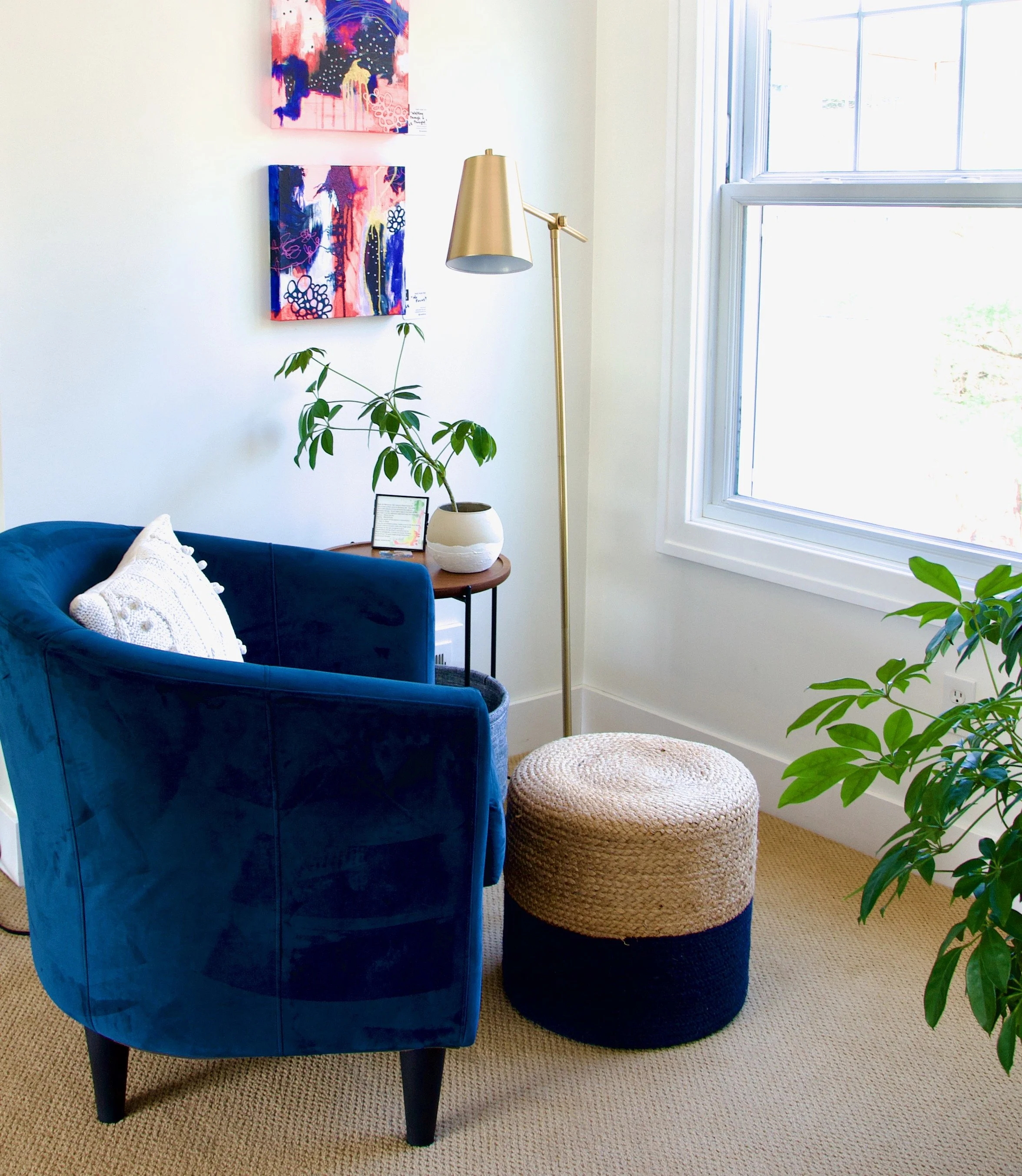 A cozy corner of a room with a plush blue velvet armchair, a beige and black woven pouf, a small side table with a potted plant, framed artwork on the wall, a tall gold floor lamp, and a window letting in natural light.