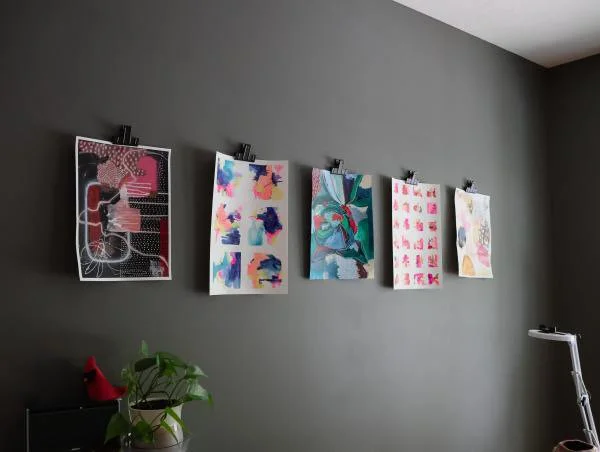 Five colorful abstract art prints hanging on a gray wall with black clips. A potted plant with large green leaves and a red decorative object are on a surface below the artwork. A white adjustable floor lamp is on the right side of the image.