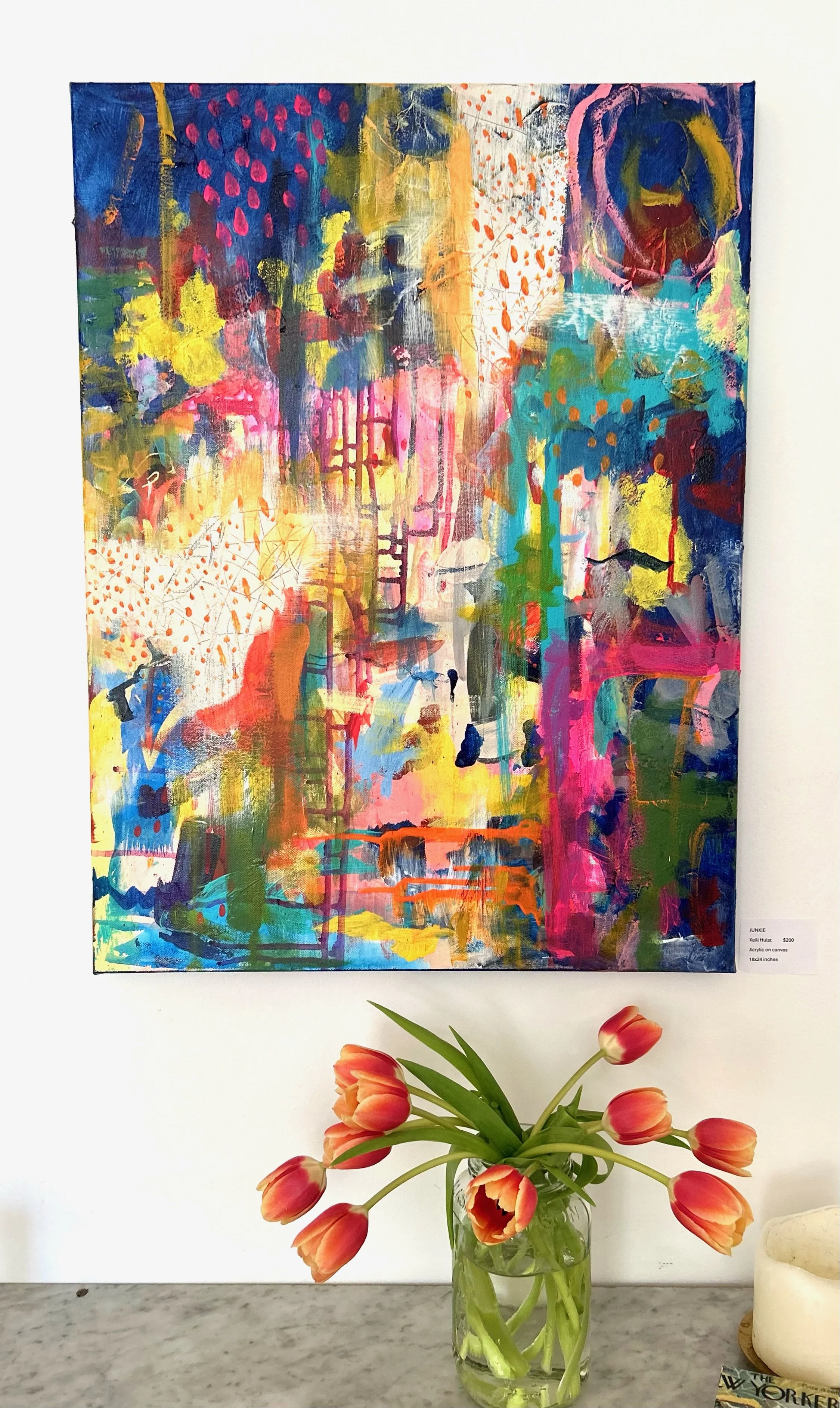 A colorful abstract painting hangs on a white wall, with a vase of pink and orange tulips in front of it on a gray surface.
