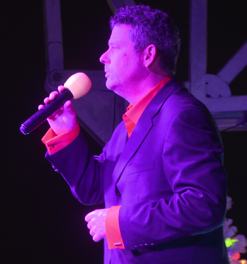 A man performing on stage, holding a microphone close to his mouth, with his eyes closed, under purple stage lighting. Tampa Bay Holiday Show