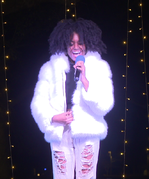 A woman with curly hair singing into a microphone on a stage decorated with string lights, wearing a white fluffy jacket and ripped jeans. Tampa Bay Holiday Show