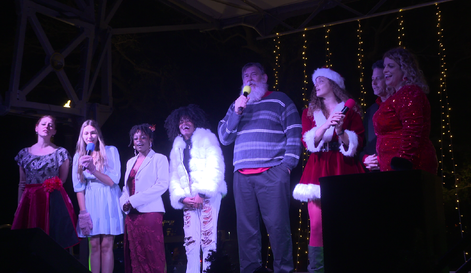 Group of people dressed in festive holiday costumes singing on stage at night, with Christmas lights in the background. Tampa Bay Holiday Show Clearwater Florida