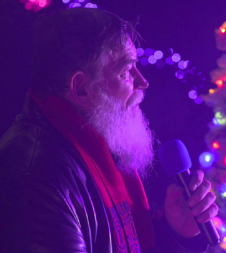 Man with a white beard and mustache holding a microphone, illuminated by purple and blue lights, with a decorated Christmas tree in the background. Tampa Bay Holiday Show Performance