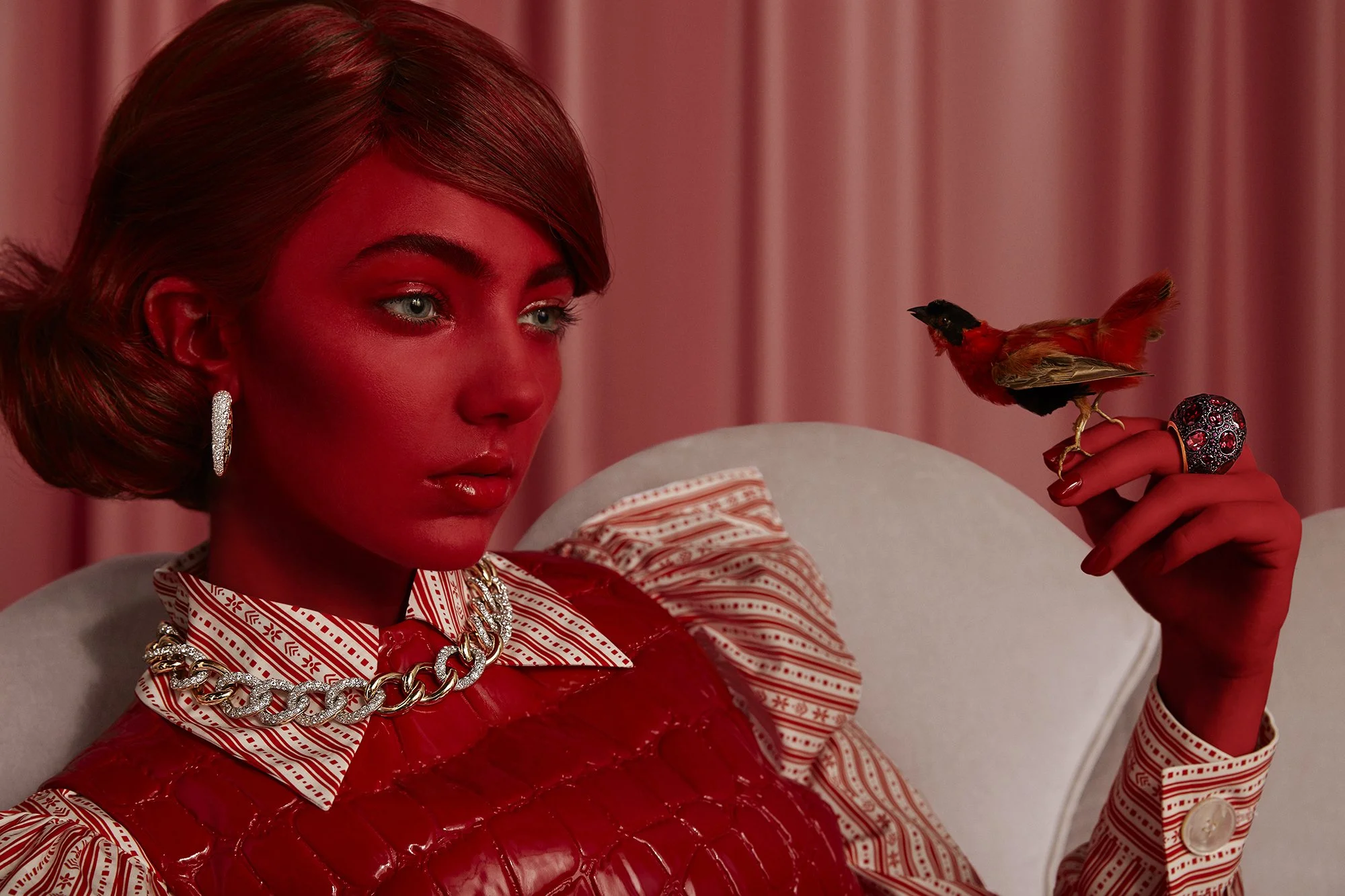 A woman with her face and hand painted entirely red, sitting on a beige chair with a pink curtain in the background. She is wearing a patterned shirt with a red leather vest, large hoop earrings, a chunky chain necklace, and a large ring. She is holding a small bird, likely a red-winged blackbird, on her finger.