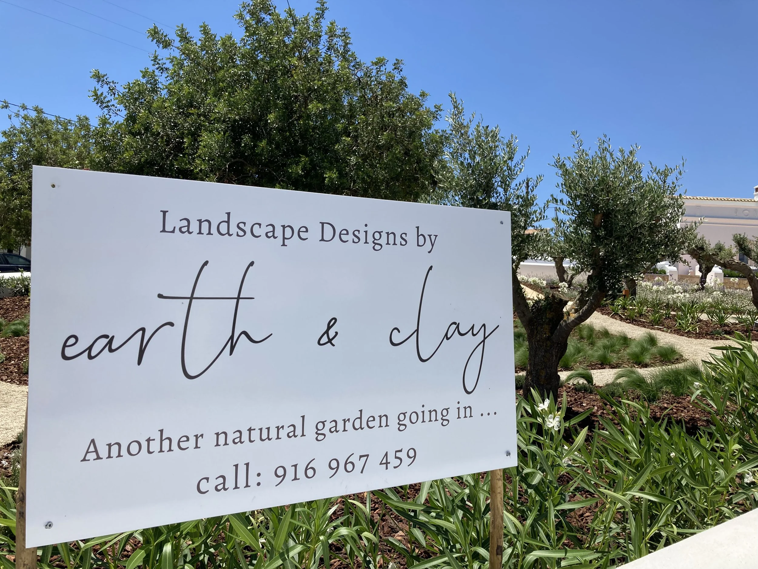 A sign in a natural garden landscape with trees and flowers, advertising landscape designs by earth & day, with a phone number, in bright sunlight under a blue sky.