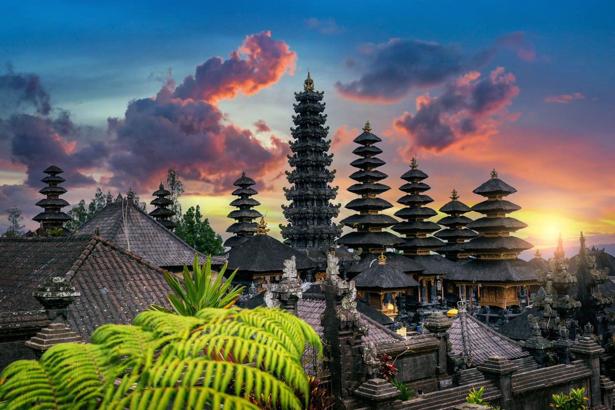 Sunset over a traditional Balinese temple complex with tiered pagoda-like structures and lush green plants in the foreground.