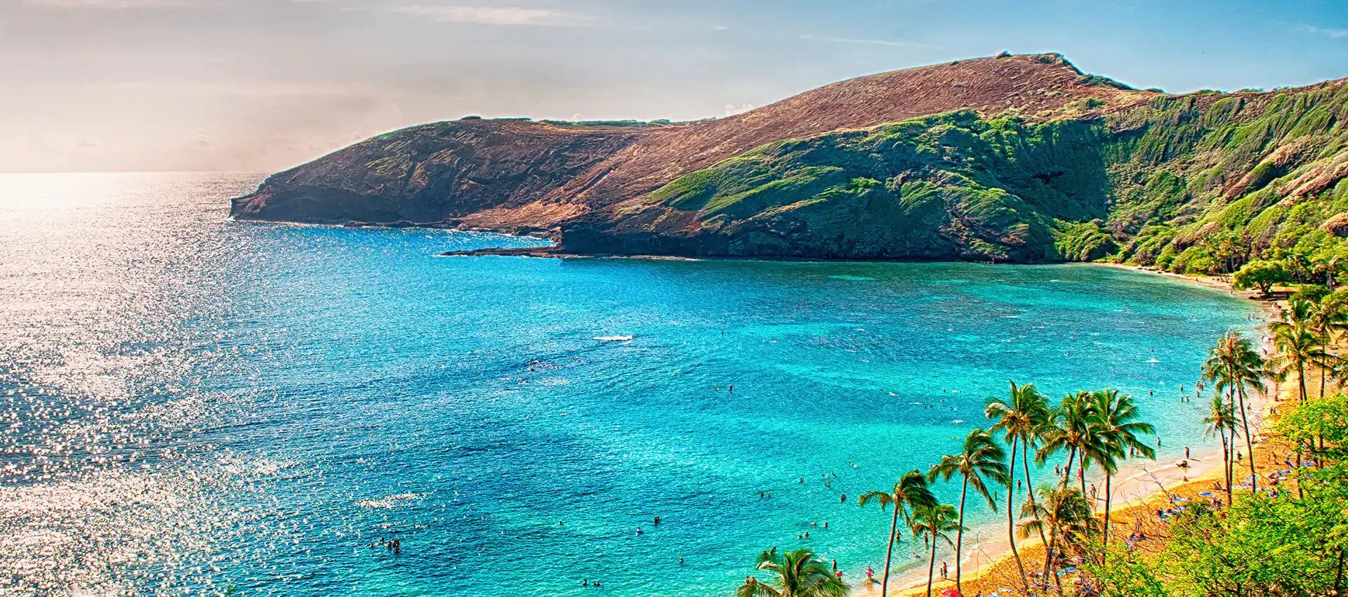 A scenic view of a coastal bay with clear turquoise waters, surrounded by lush green hills and a sandy beach with palm trees and people enjoying the beach.