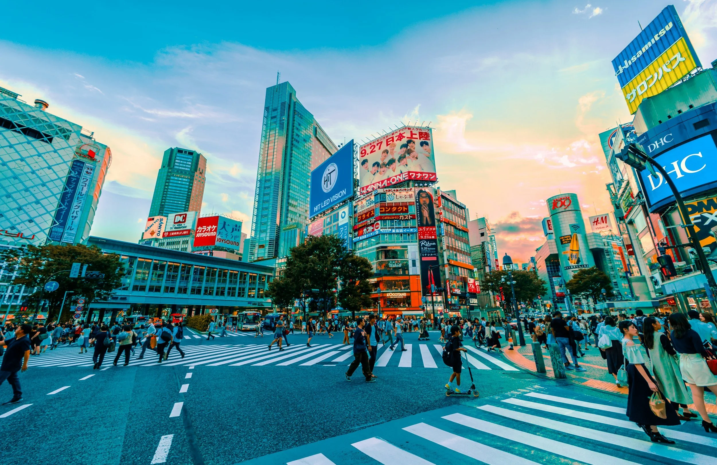 Crowded city street at dusk with tall buildings, billboards, and a crosswalk filled with pedestrians.