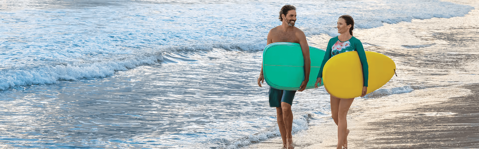 A man and woman walking on the beach carrying surfboards, smiling at each other with ocean waves in the background.