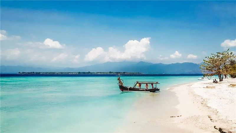 A tropical beach with white sand, turquoise water, a boat near the shore, and a tree on the right side. Mountains and clouds are visible in the background.