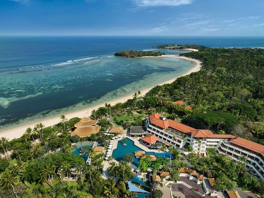 Aerial view of a tropical resort with pools, surrounded by lush greenery, next to a sandy beach and calm ocean waters
