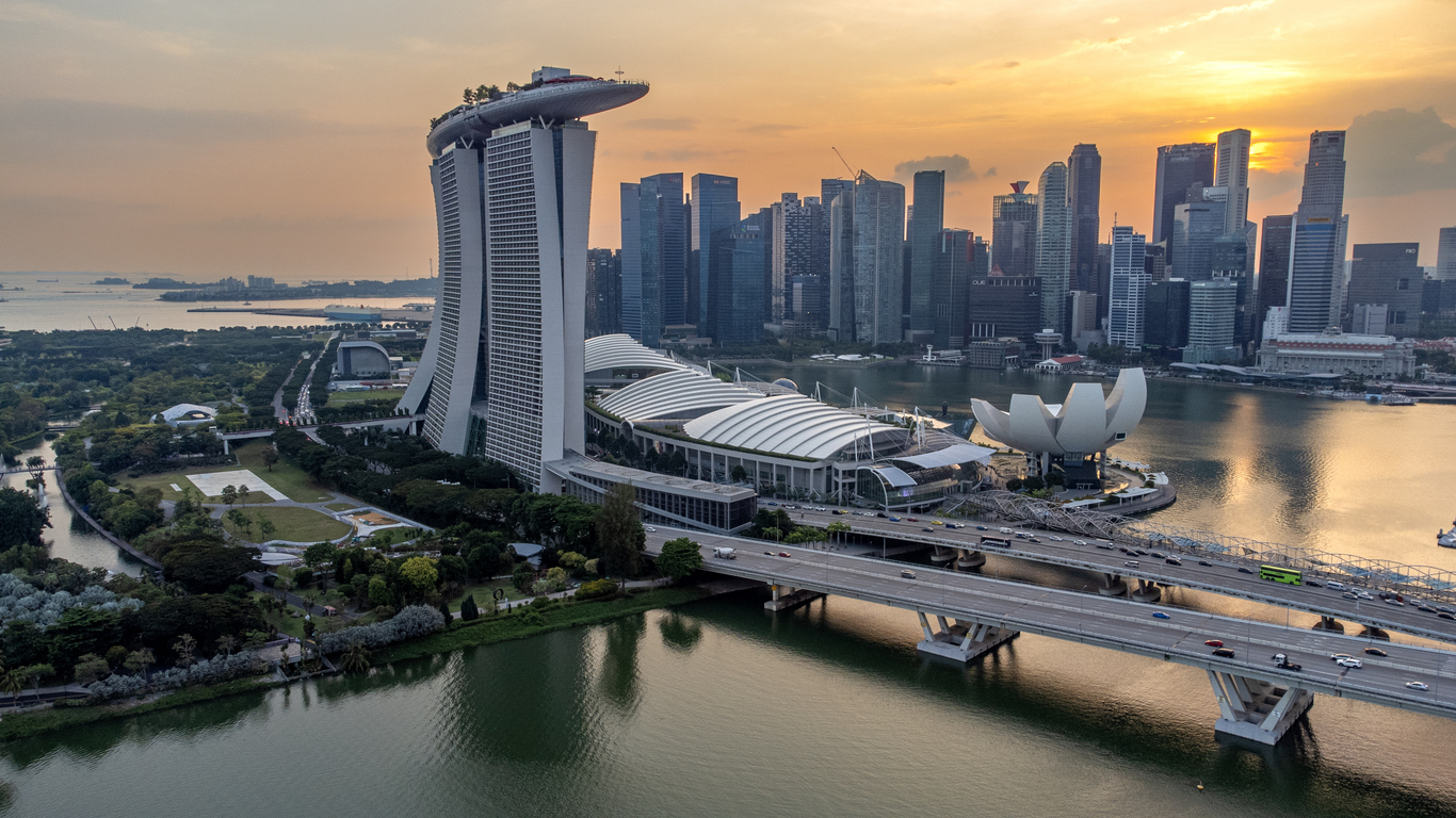 A city skyline at sunset featuring the Marina Bay Sands hotel with its boat-shaped top, nearby Museum of Art Science with a lotus flower design, and numerous skyscrapers in the background, along with a bridge over a body of water in the foreground.