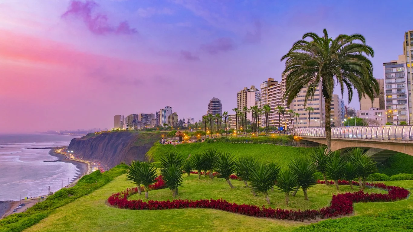 Cityscape with high-rise buildings along a coastline at sunset, featuring palm trees, lush greenery, and a cliff overlooking the ocean.