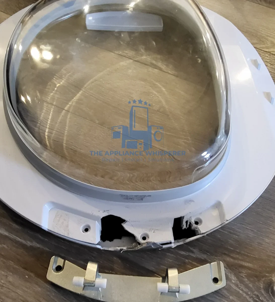 Broken or damaged component of an appliance, possibly a washing machine or dryer, with a silver tinted transparent cover and some white plastic and beige parts on a wooden surface.