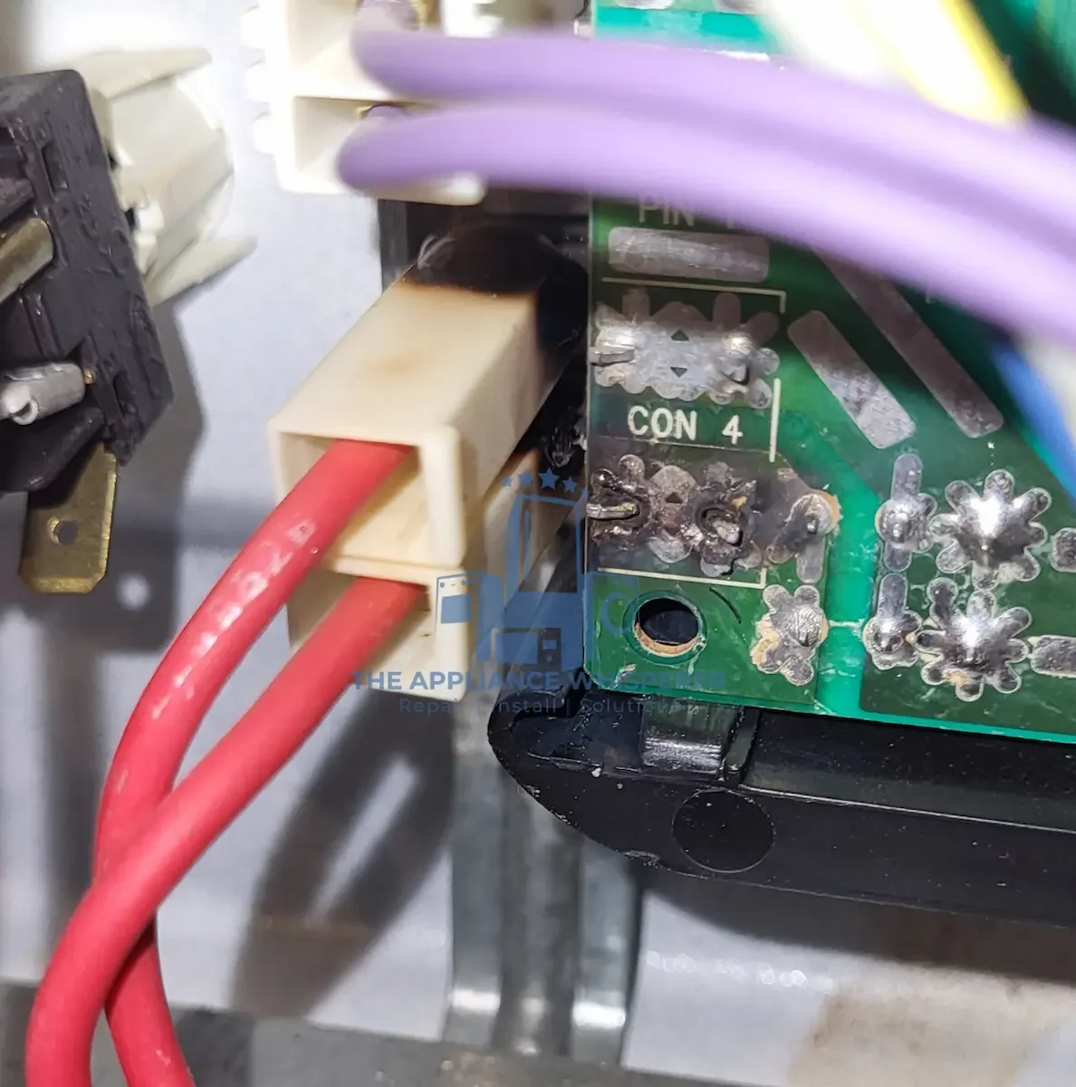 Close-up of an electrical circuit board with red wires connected to a white plastic connector, and soldered connections on the green circuit board, with other electrical components nearby.