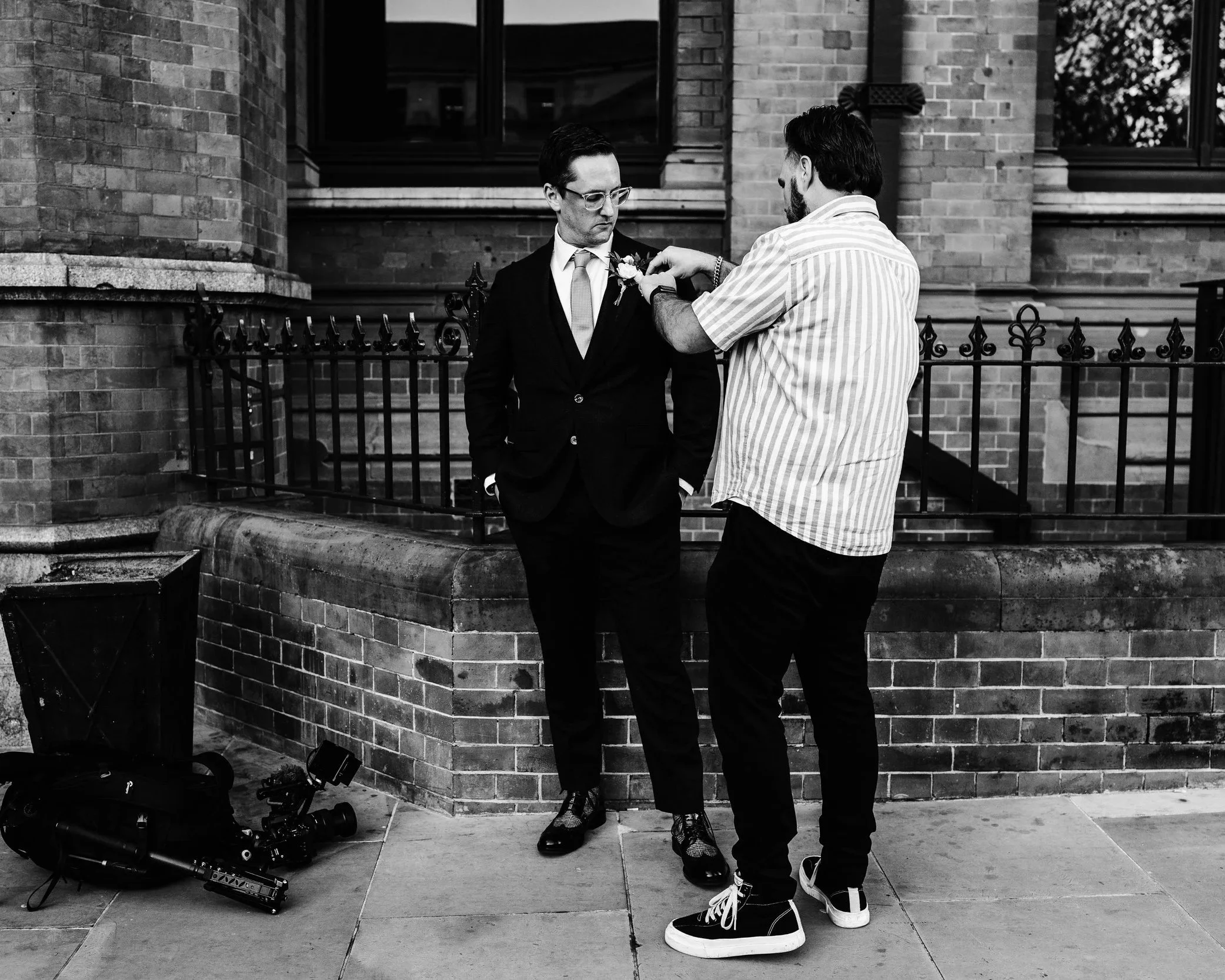 Two men standing outside a brick building. One man is dressed in a suit and tie with glasses, while the other man is wearing a striped shirt. The man in the striped shirt is pinning a boutonniere onto the lapel of the suited man. There is a camera and tripod on the ground nearby.