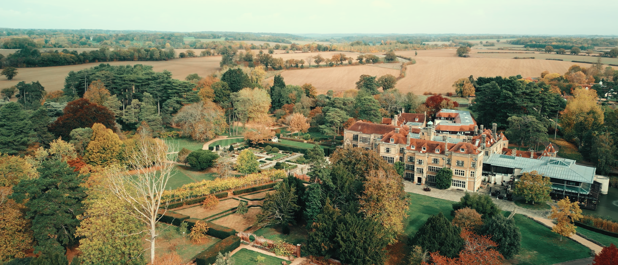Aerial view of a large historic mansion surrounded by beautifully maintained gardens and numerous trees with autumn foliage. The mansion features red tile roofs and large windows, with fields and forests stretching into the distance.