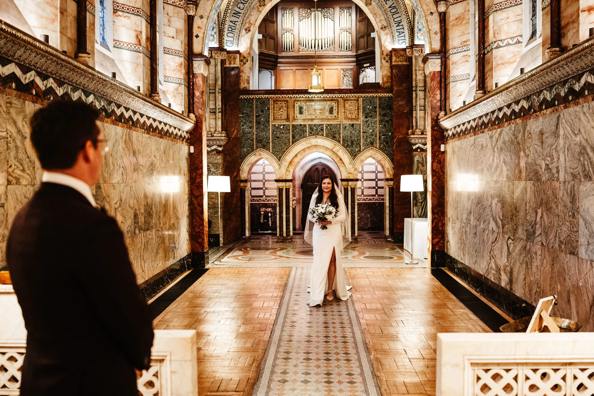 A bride in a white gown holding a bouquet walks down the aisle in a decorated church with gold accents, while a man in a tuxedo stands at the front watching her.