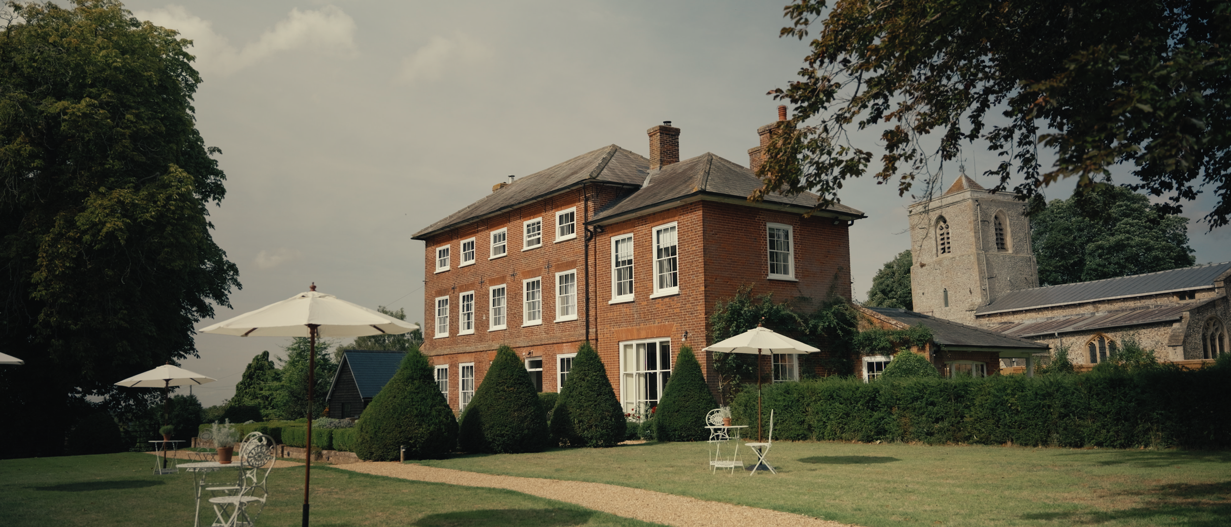 Sandon Manor with white windows, surrounded by trimmed bushes and garden furniture with umbrellas, and a stone church in the background under a partly cloudy sky.