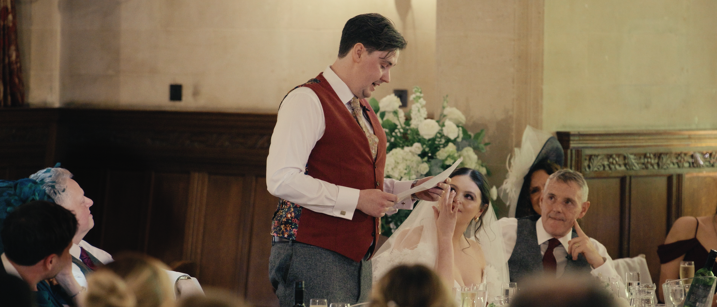 Man in a red vest giving a speech at a wedding reception, with a bride and guests listening.