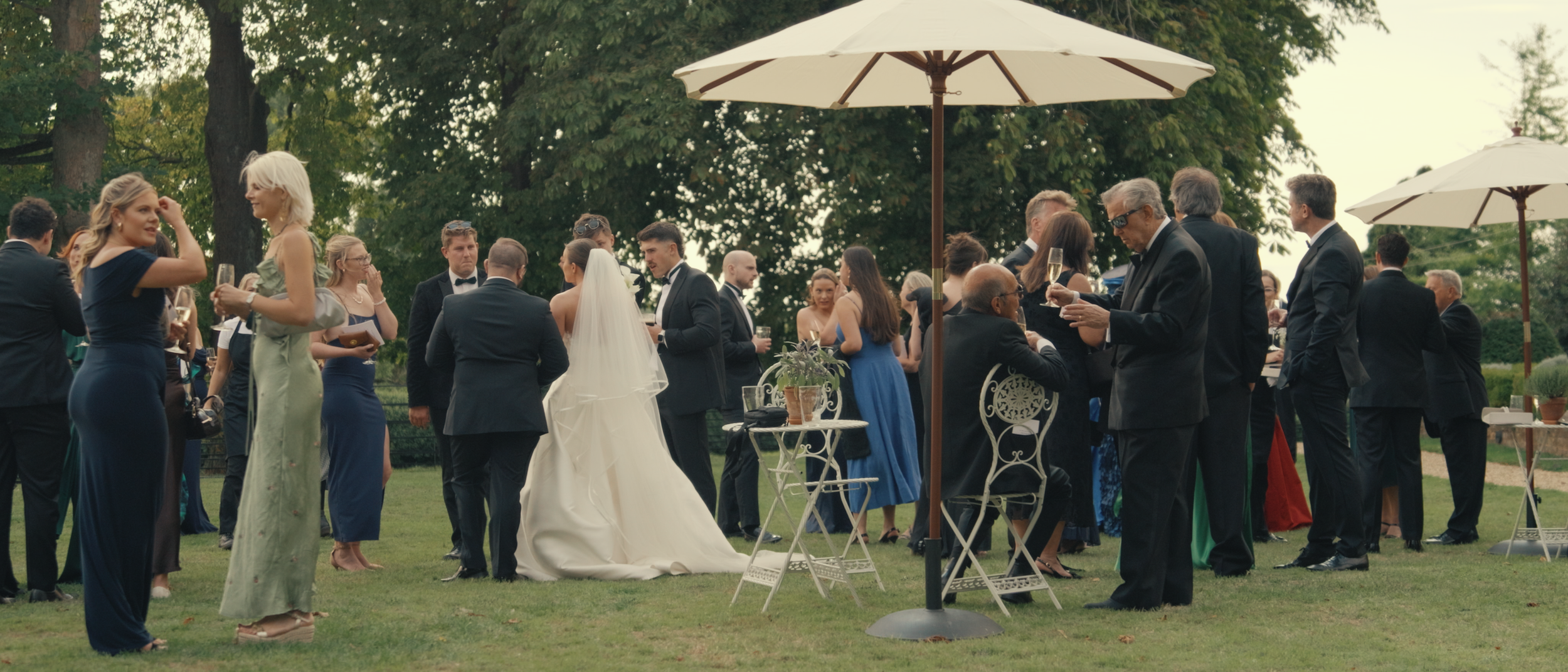 Guests at a wedding reception outdoors, dressed in formal attire, socializing under umbrellas on the lawn with trees in the background.