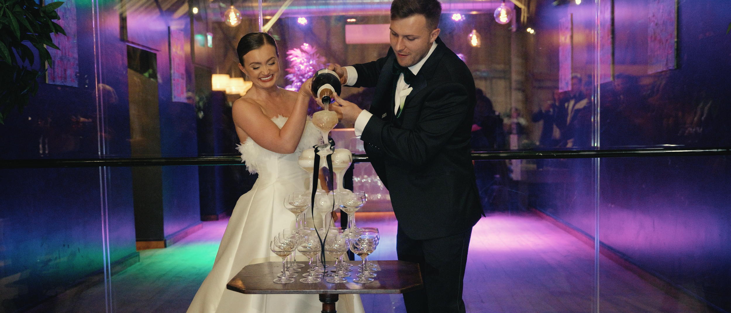 A bride and groom in wedding attire pour champagne into glasses on a table during their celebration.