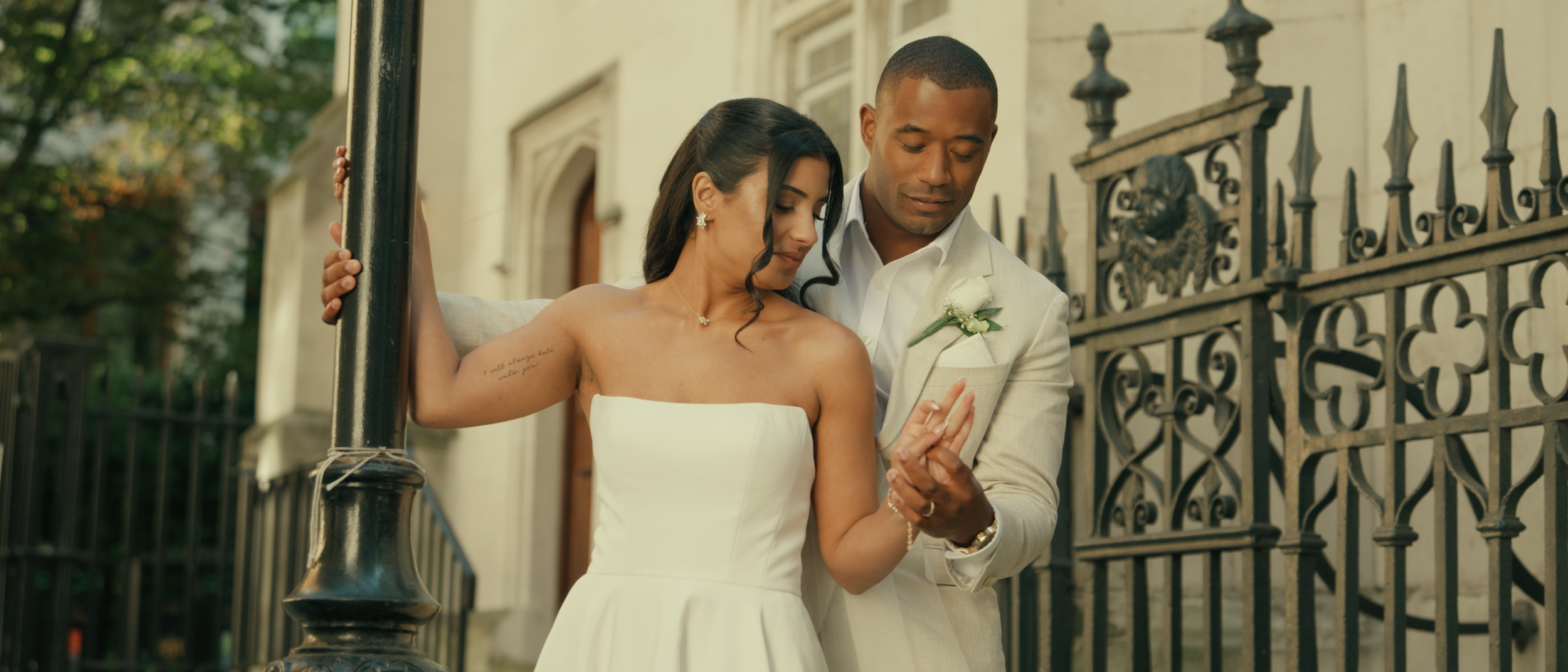 A newlywed couple, a woman in a strapless white dress and a man in a light-colored suit, share a tender moment by a wrought-iron fence outside a historic building.