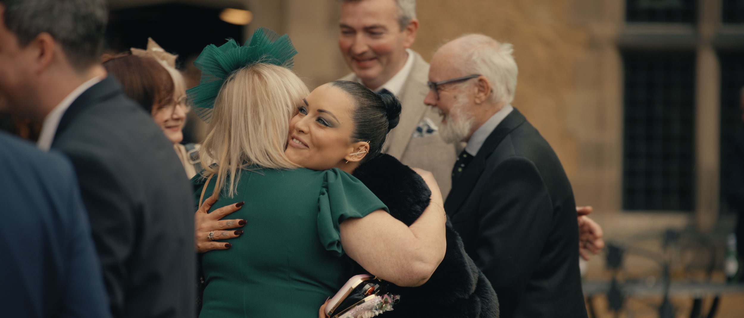 A woman with dark hair and makeup hugging an older blonde woman with a teal headband and green dress at a social gathering.