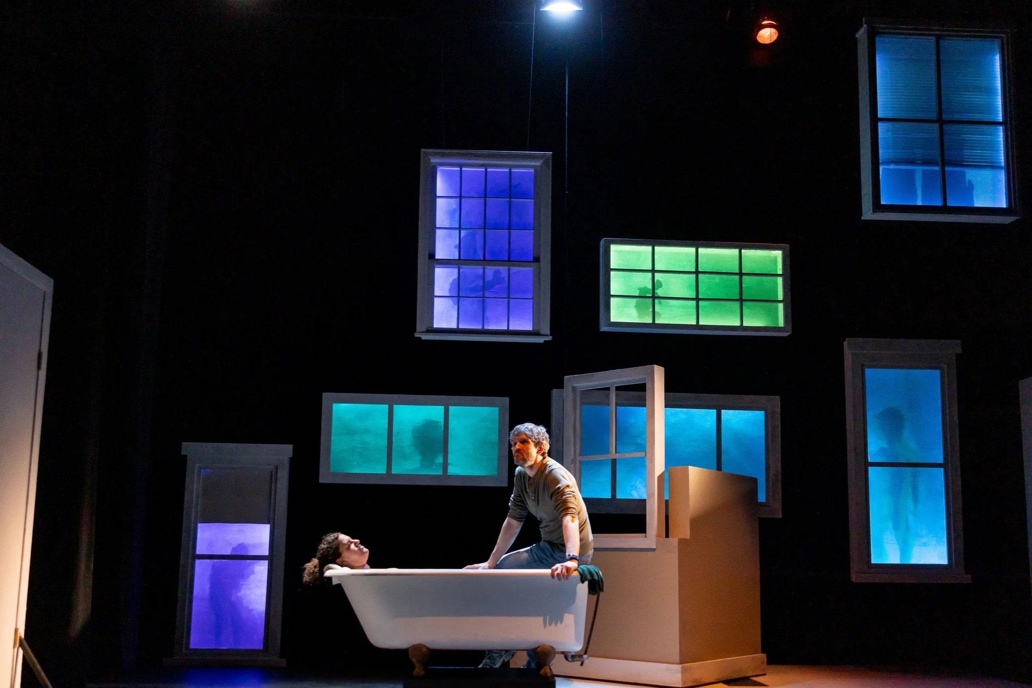 Man sits on edge of tub with woman soaking, in front of illuminated cool toned windows.