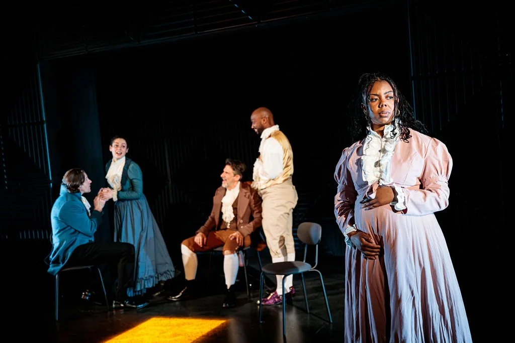 A pregnant woman in 1800s dress stands in the foreground, while her friends laugh in the background