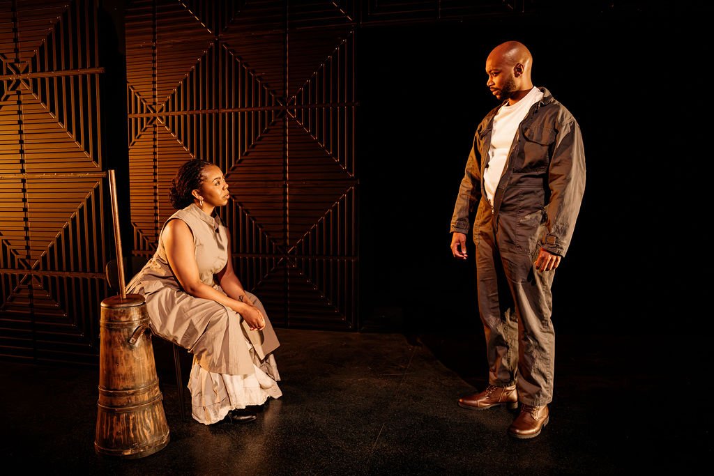 A woman sits with a butter churn looking at a man in coveralls