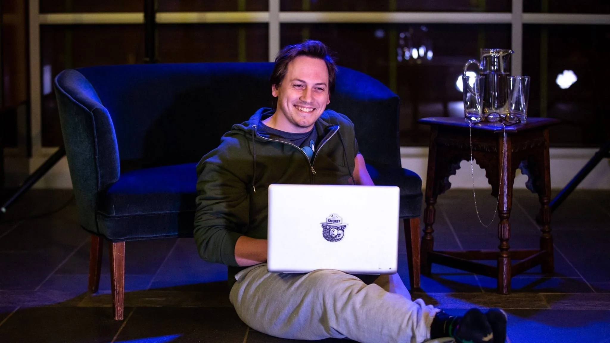 Smiling man in sweats sits on ground with laptop.