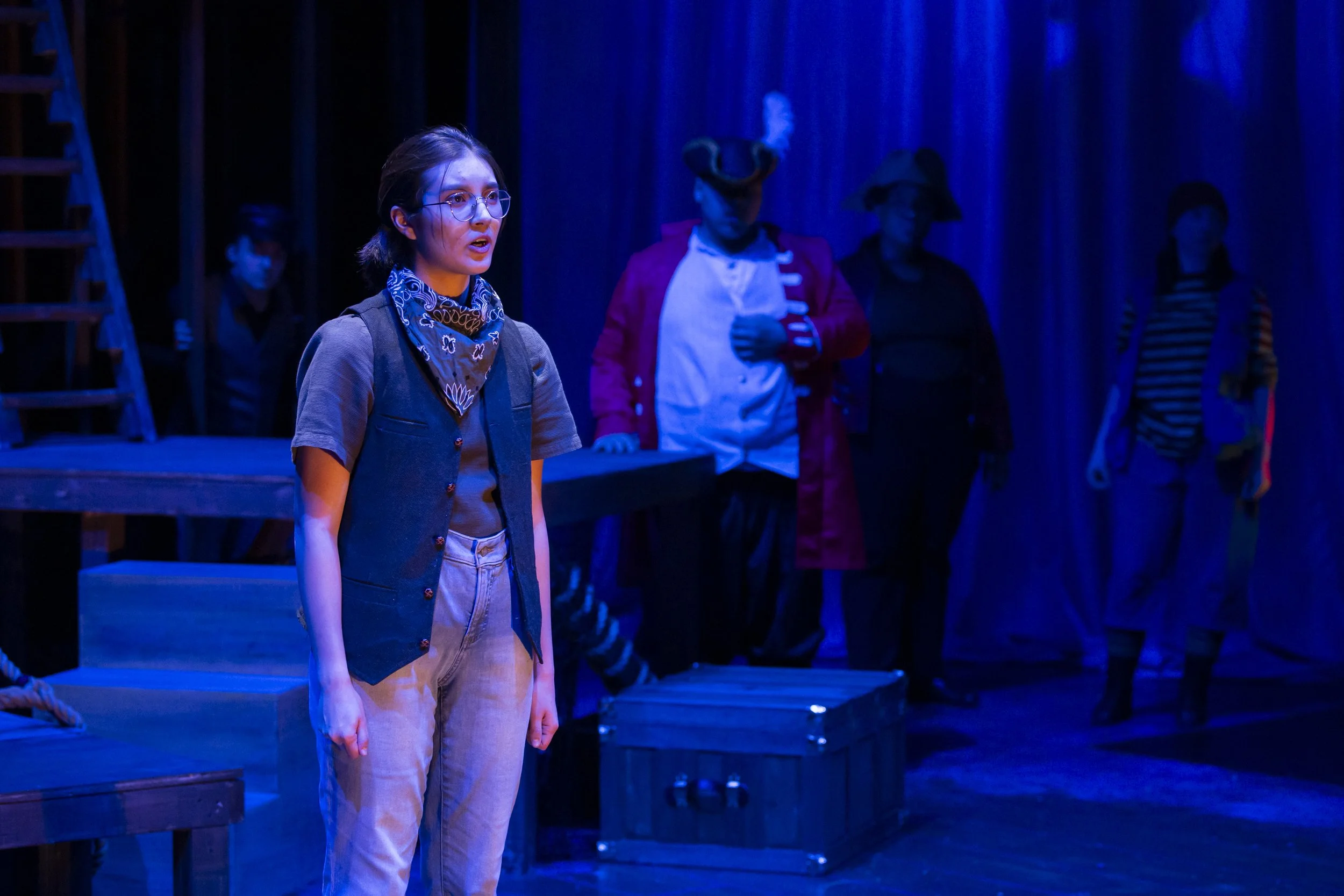 Young boy stands wistfully in dramatic blue light, as cast looks on.