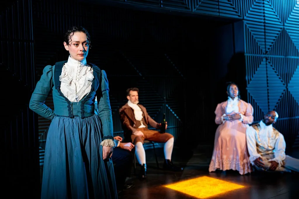 Mary Shelley stands in the foreground, while others sit in the background, against a futuristic set