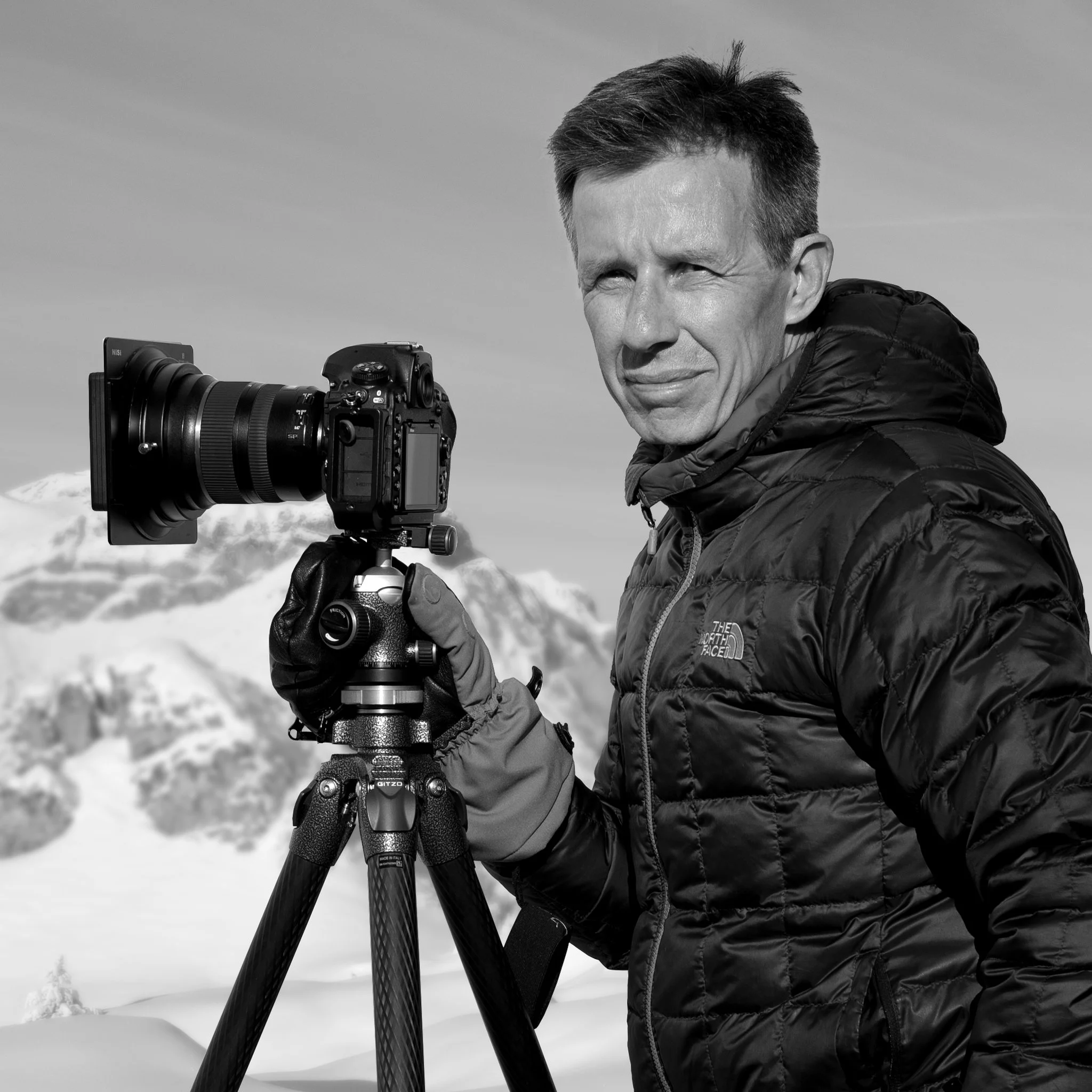 René Schädler stands with a camera on a tripod in front of a snow-covered mountain landscape.